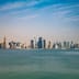 A bright, panoramic view of the modern Doha skyline rising across the water under a clear blue sky.