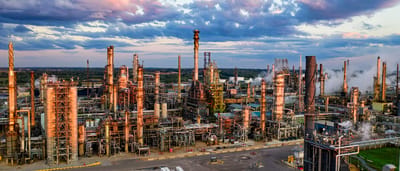 An expansive aerial view of an oil refinery complex with towering smokestacks and pipelines illuminated under a dramatic sunset sky.
