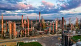 An expansive aerial view of an oil refinery complex with towering smokestacks and pipelines illuminated under a dramatic sunset sky.