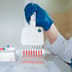 A scientist wearing blue gloves uses a multichannel pipette to dispense red liquid into a microplate in a modern laboratory.