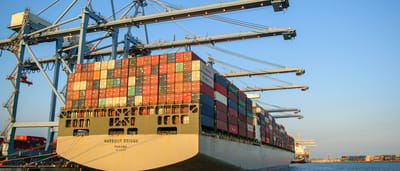 A massive, fully loaded cargo ship docked at an industrial port under a clear blue sky.