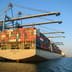 A massive, fully loaded cargo ship docked at an industrial port under a clear blue sky.