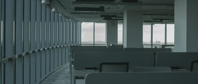 A quiet, empty modern office space with rows of vacant desks and large windows facing a grey sky.