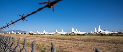 A row of parked military aircraft sitting idle in a desert boneyard, viewed through a barbed wire fence.
