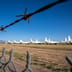 A row of parked military aircraft sitting idle in a desert boneyard, viewed through a barbed wire fence.